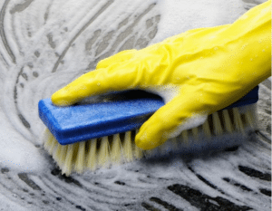 Hand with yellow glove scrubbing garage floor with brush and cleaning solution.